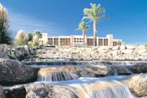 Valle del Este Hotel with cascading waterfall feature in the foreground, surrounded by rocky landscape and palm trees under a clear blue sky.