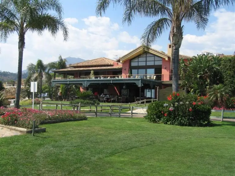 Clubhouse at Los Arqueros Golf Club surrounded by lush greenery, palm trees, and landscaped gardens on a sunny day.