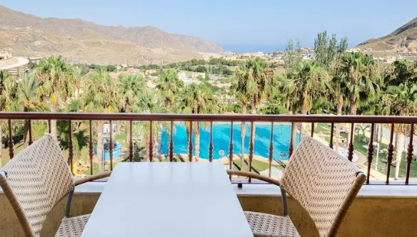 View from a balcony at Hotel La Envia Spa and Golf Resort in Almeria, overlooking a large swimming pool surrounded by palm trees, with a backdrop of rolling hills and the distant sea.