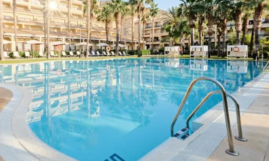 Large outdoor swimming pool at Hotel La Envia Spa and Golf Resort in Almeria, surrounded by sun loungers, palm trees, and the hotel's multi-story building in the background.