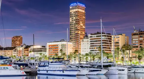 Nighttime view of the Alicante Gran Sol hotel towering over the marina, with yachts and city lights reflecting on the water.