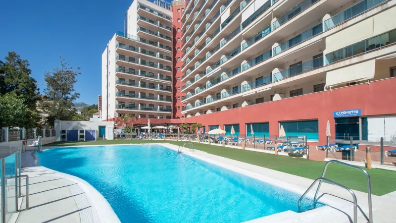 Outdoor swimming pool at Hotel Principe Apartments, surrounded by sun loungers and umbrellas, with a multi-story building featuring balconies in the background under a clear blue sky.